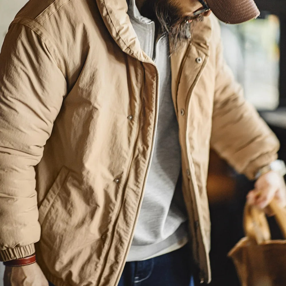 A man wearing a casual, beige, windbraker jacket, and is looking down into the floor